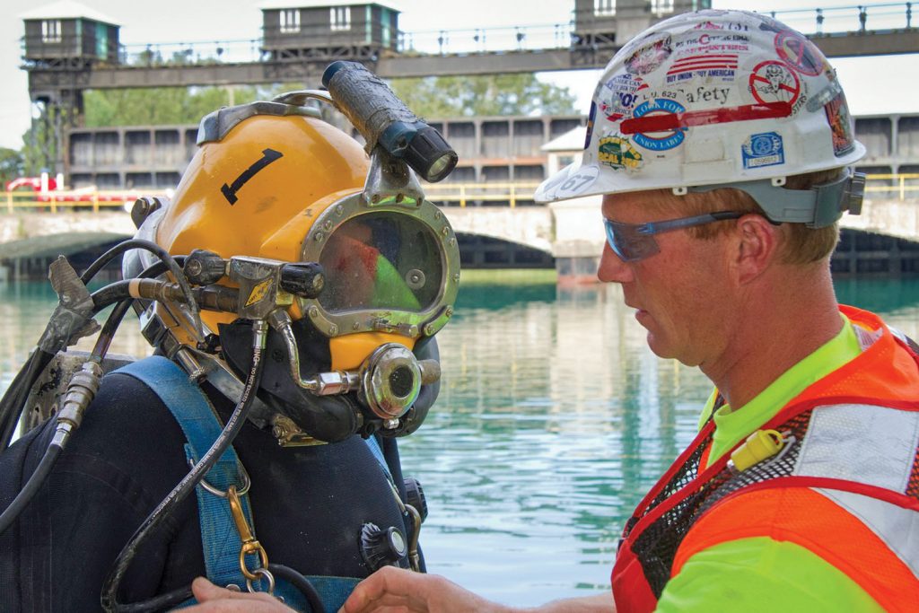 A dive tender, in hard hat, double checks the diver’s equipment prior to the dive. The tender will keep his attention on the umbilicals and monitoring equipment the entire time the diver is underwater. (Photo courtesy of J.F. Brennan Inc.)