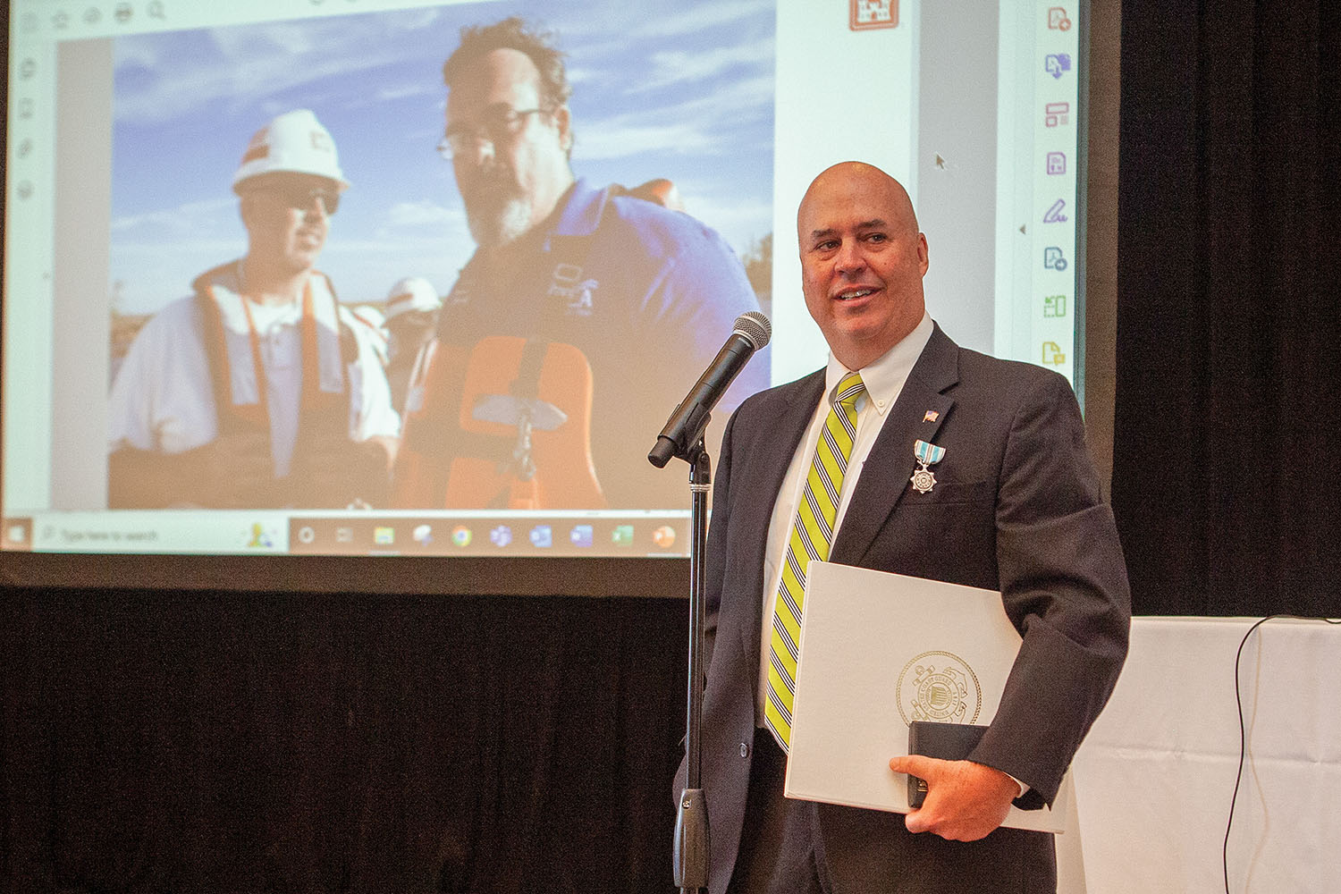 Vic Landry, operations manager of the GIWW, speaks to the GICA crowd after receiving the Coast Guard Meritorious Public Service Award. Landry and Paul Dittman, GICA president, are pictured together on the screen during Hurricane Ida recovery operations. (Photo by Frank McCormack)